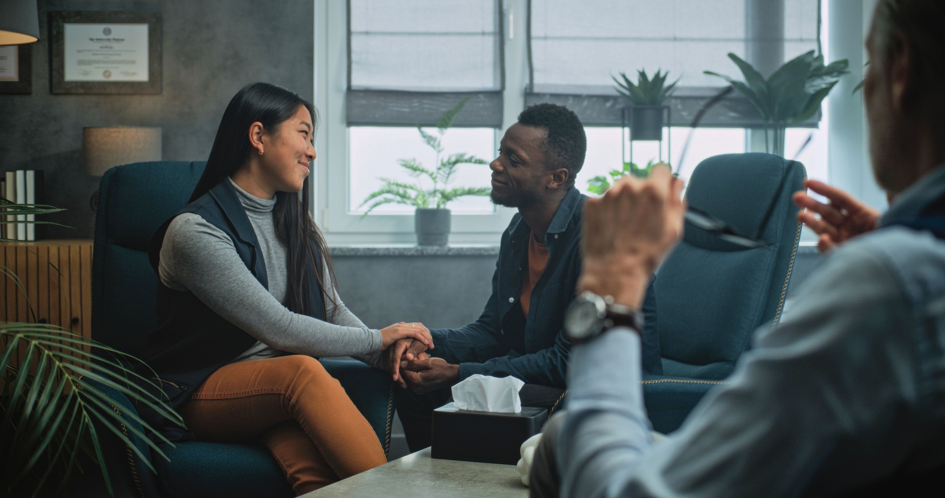 Happy multiethnic couple sit on chairs hand in hand on couples therapy